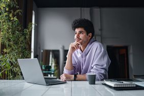 A man sits with a laptop at a table, looking into the distance and pondering.