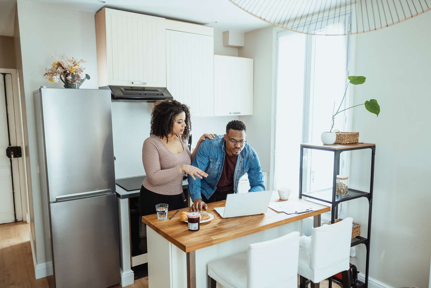 A couple standing together in a well-lit kitchen discuss something on a laptop screen.