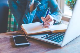 Student at desk, writing in journal on top of open laptop