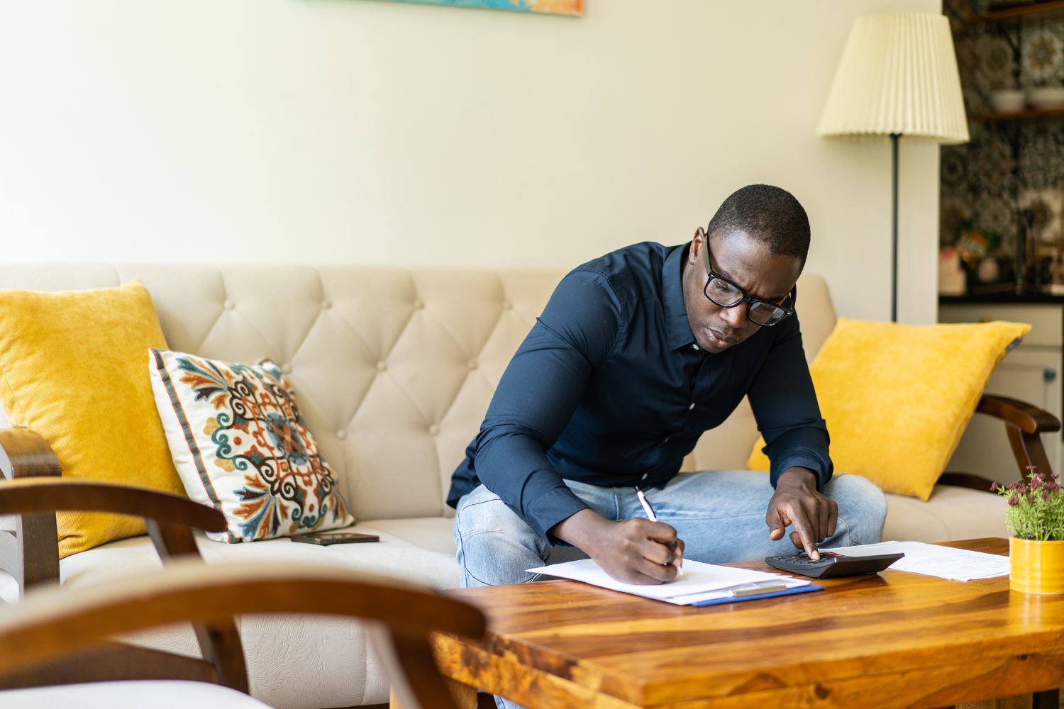A man leans over a coffee table and fills out a form attached to a clipboard. Thereâs a calculator on the table near the form