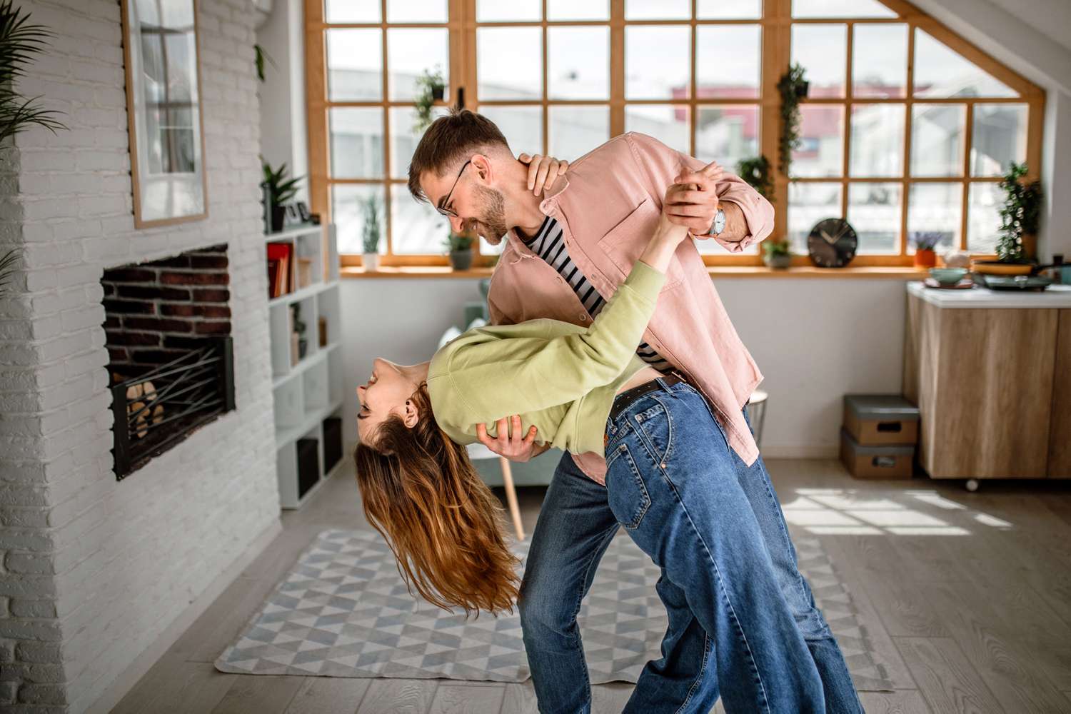 Young Millennials couple with high income dancing in their spacious, bright living room with a fireplace and big window.
