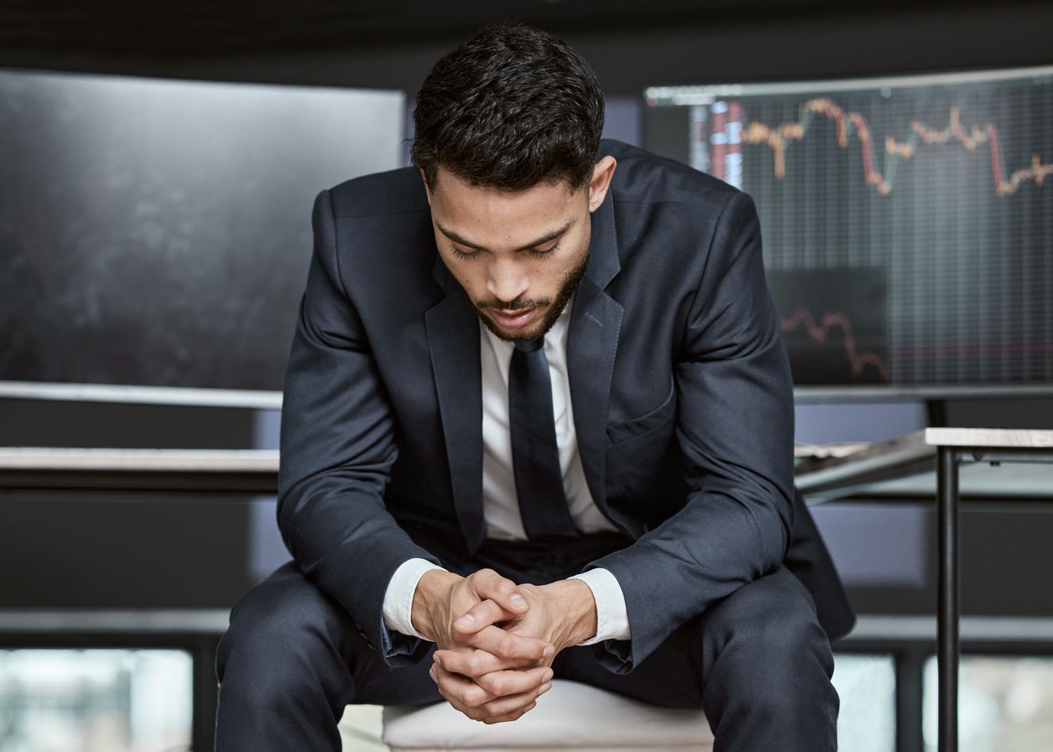 A man in a business suit sitting with his head down, stock market monitors in the background
