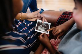 A close-up image of a group of people sitting together, focusing on a person holding an ultrasound photo. Another person is pointing at the image, which appears to be a baby in the womb. The scene conveys a sense of joy, connection, and anticipation, with warm tones and cozy clothing, including stripes and plaid patterns.
