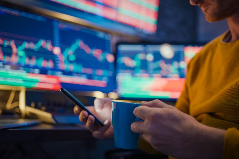 A person holding a mug and holding a mobile phone in front of a bank of computer montiors