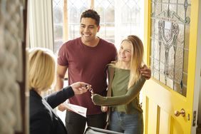 A smiling couple stands at the doorway of their new home, receiving a set of keys from a real estate agent. The bright yellow door and stained-glass window add warmth to the welcoming scene.
