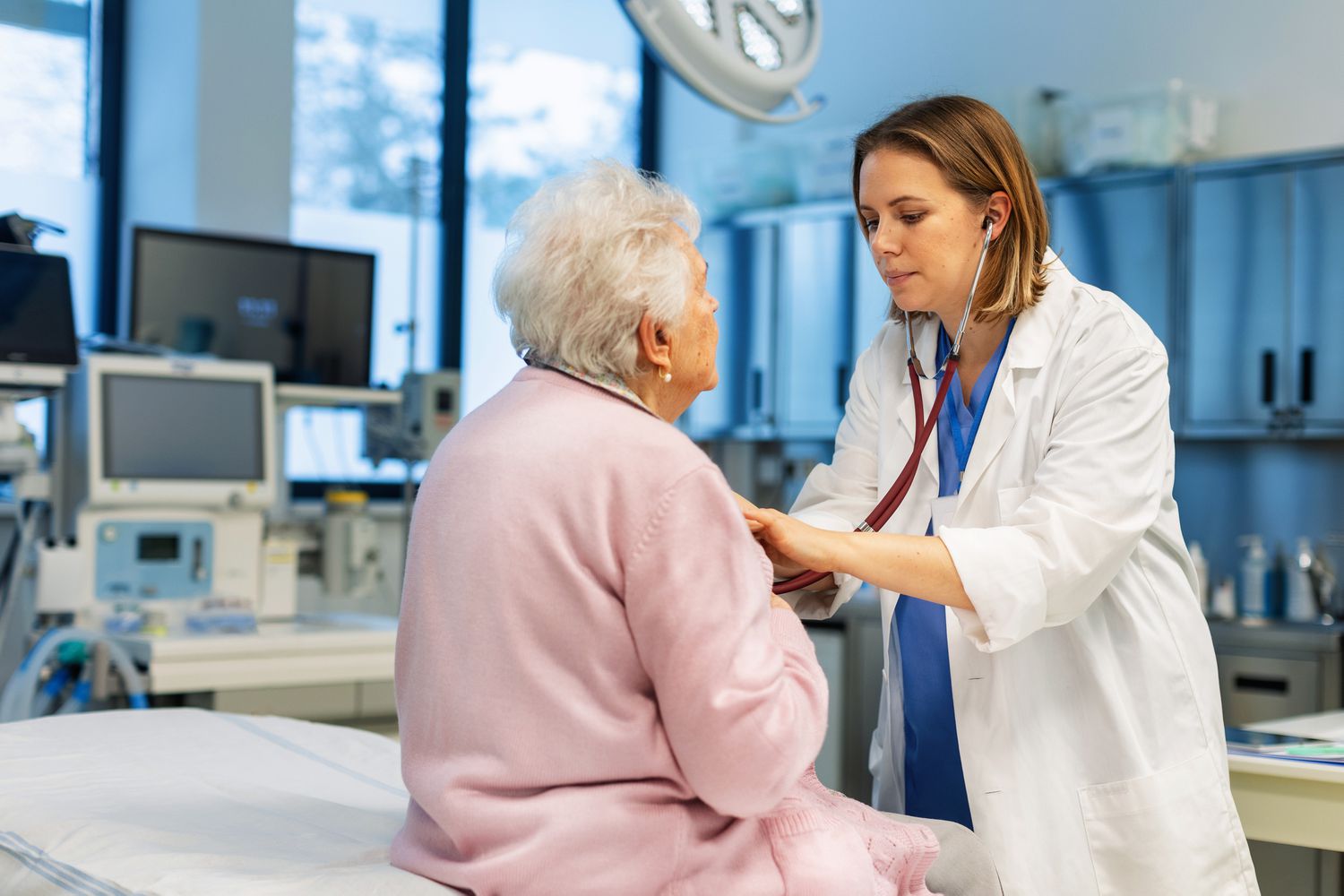 A doctor uses a stethoscope to listen to the chest of a mature adult sitting on an exam table.