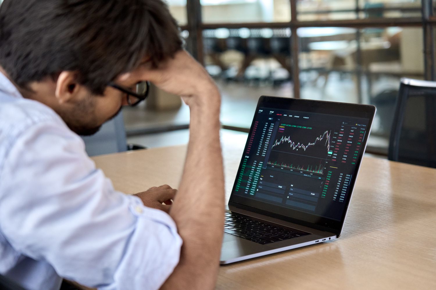 An investor sitting at a desk with their head in their hands looking unconfidently in the stock trades on their computer screen.