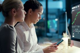 Two women looking at a smartphone and computer screen with financial market information on them.