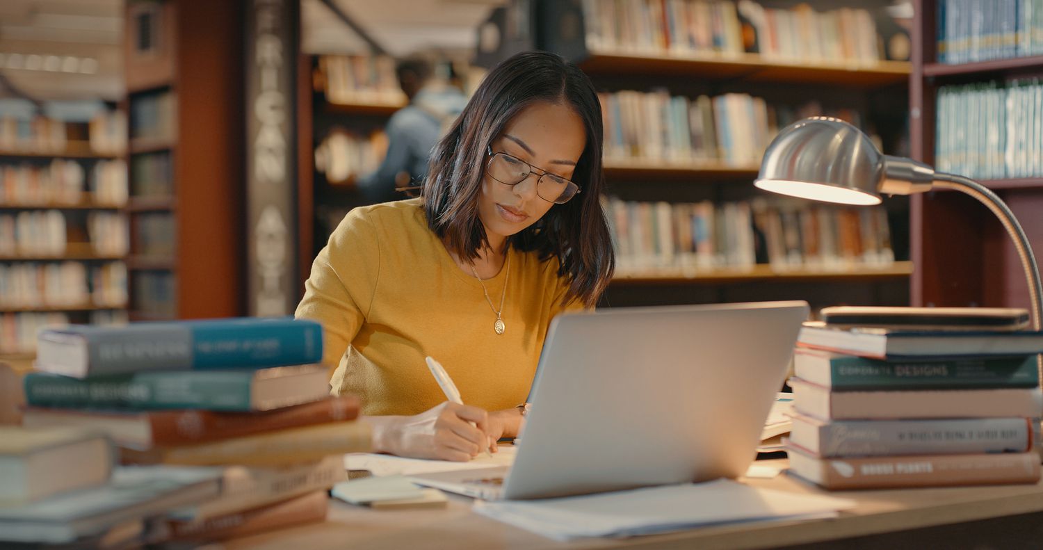 Young lady using a laptop to do research on the internet while sitting in a library surrounded by books