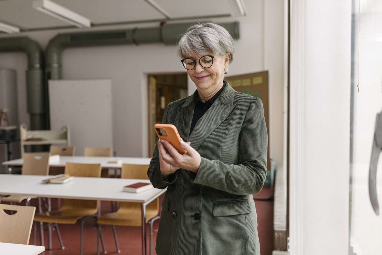 A teacher smiling while using her smartphone and standing in an empty classroom.
