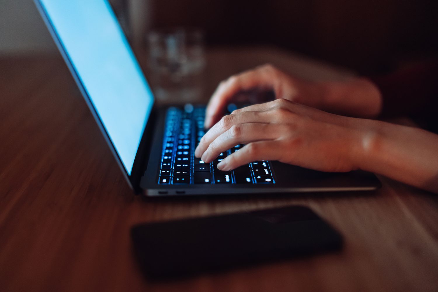 Close up of hands typing on computer keyboard while sitting on desk in the evening, with smartphone by the side