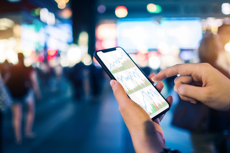 A stock trading app user reviews charts and balances with a mobile app in a crowded shopping and dining district.