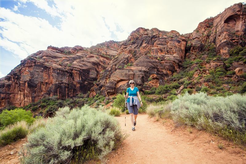 A retired woman is enjoying a walk along a scenic trail with vibrant red rock cliffs in the background.