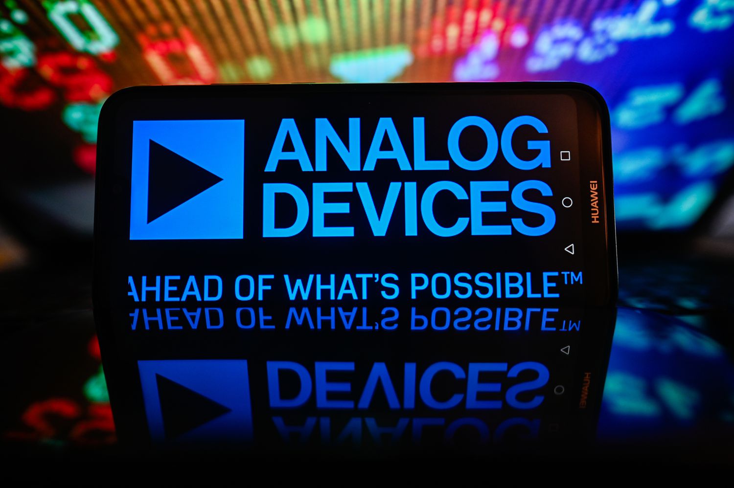 An Analog Devices logo is displayed on a smartphone with stock market percentages in the background