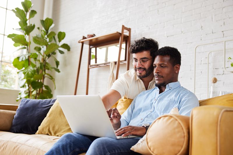 A man sits on a couch with a laptop while another man stands behind him looking over his shoulder and pointing at the laptop