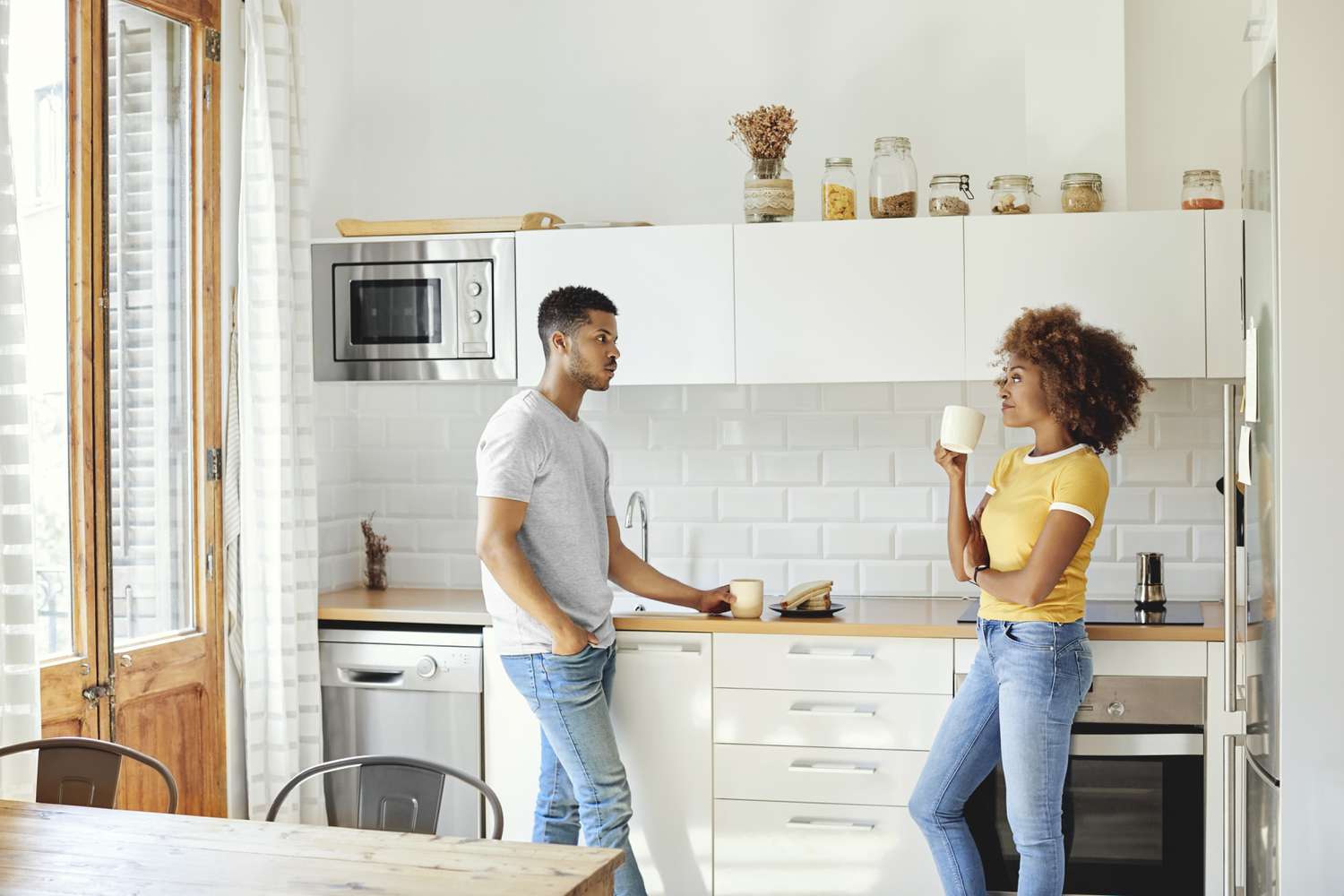 A man and a woman stand in a kitchen holding mugs and talking