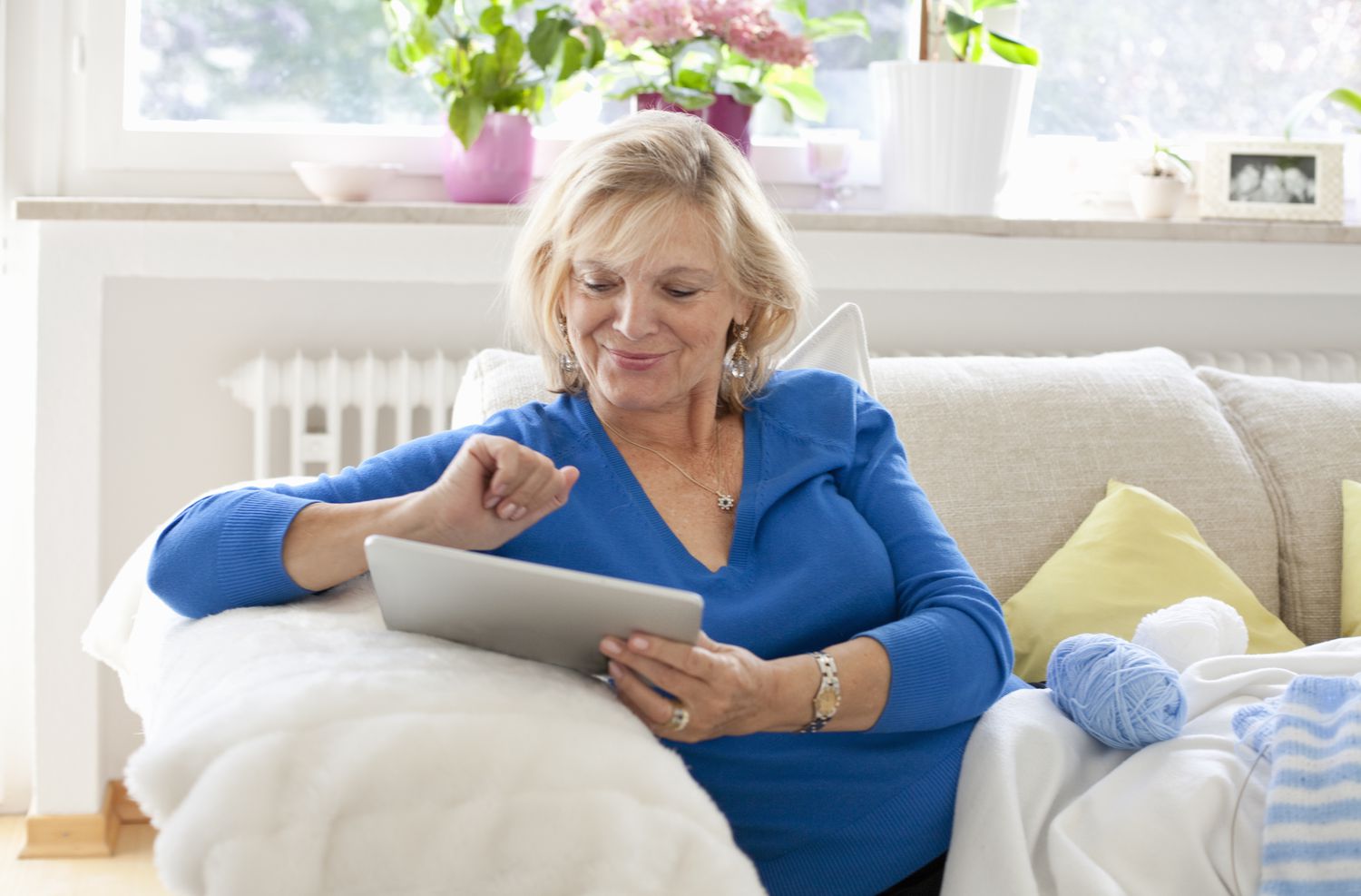 Older woman sitting on her living room couch and smiling as she looks at something on her tablet.