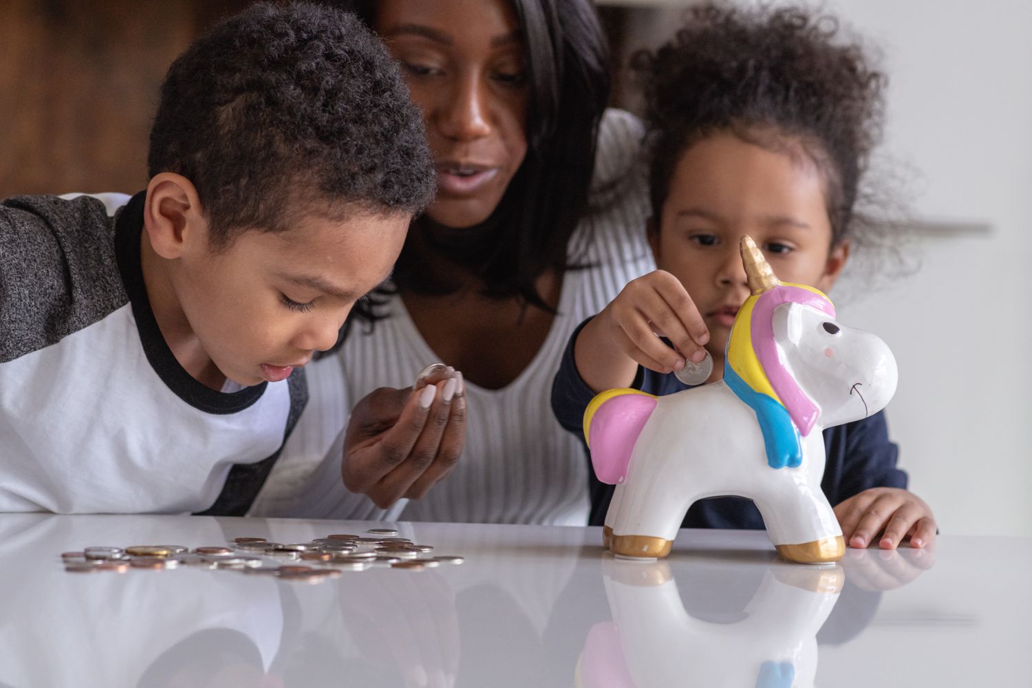 A mother holds a coin, with other coins on a table, as you son looks at a pile of coins and a daughter puts a quarter into a unicorn piggy bank.