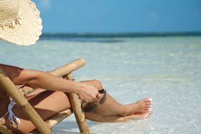 Woman sitting in a chair at the beach, wearing a sun hat, holding sunglasses, and with her feet in the water