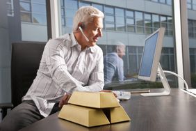 Man sitting at a table working on a computer with gold bars stacked beside him.