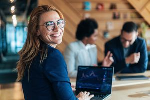 A woman smiling and working on a laptop, with two people discussing in the background
