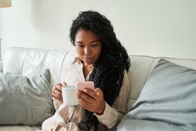 Young woman sitting on her sofa with a cup of tea, looking seriously at news on her smartphone