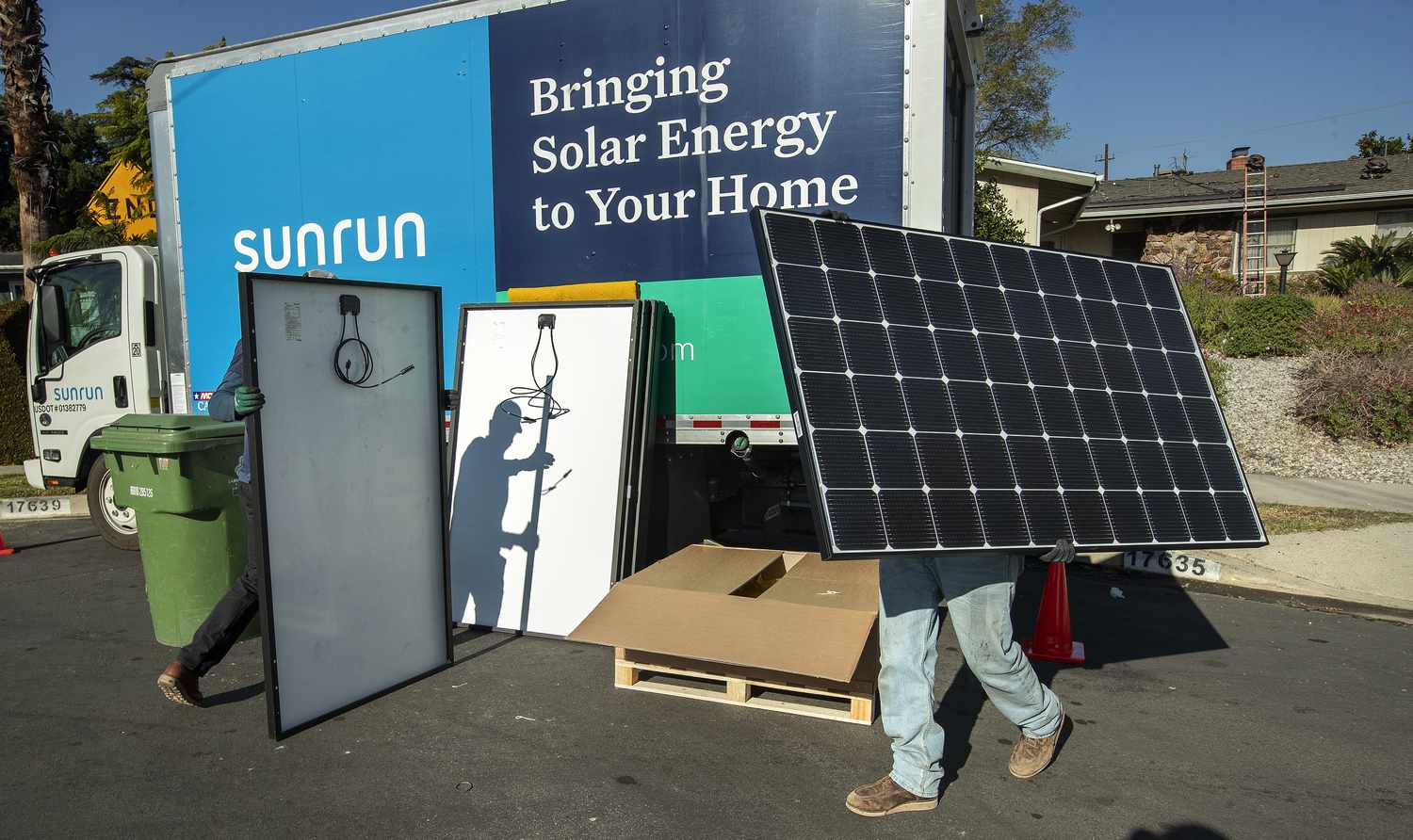 Two employees of Sunrun carry solar panels to be installed on the roof of a home, walking past a truck with a Sunrun logo.