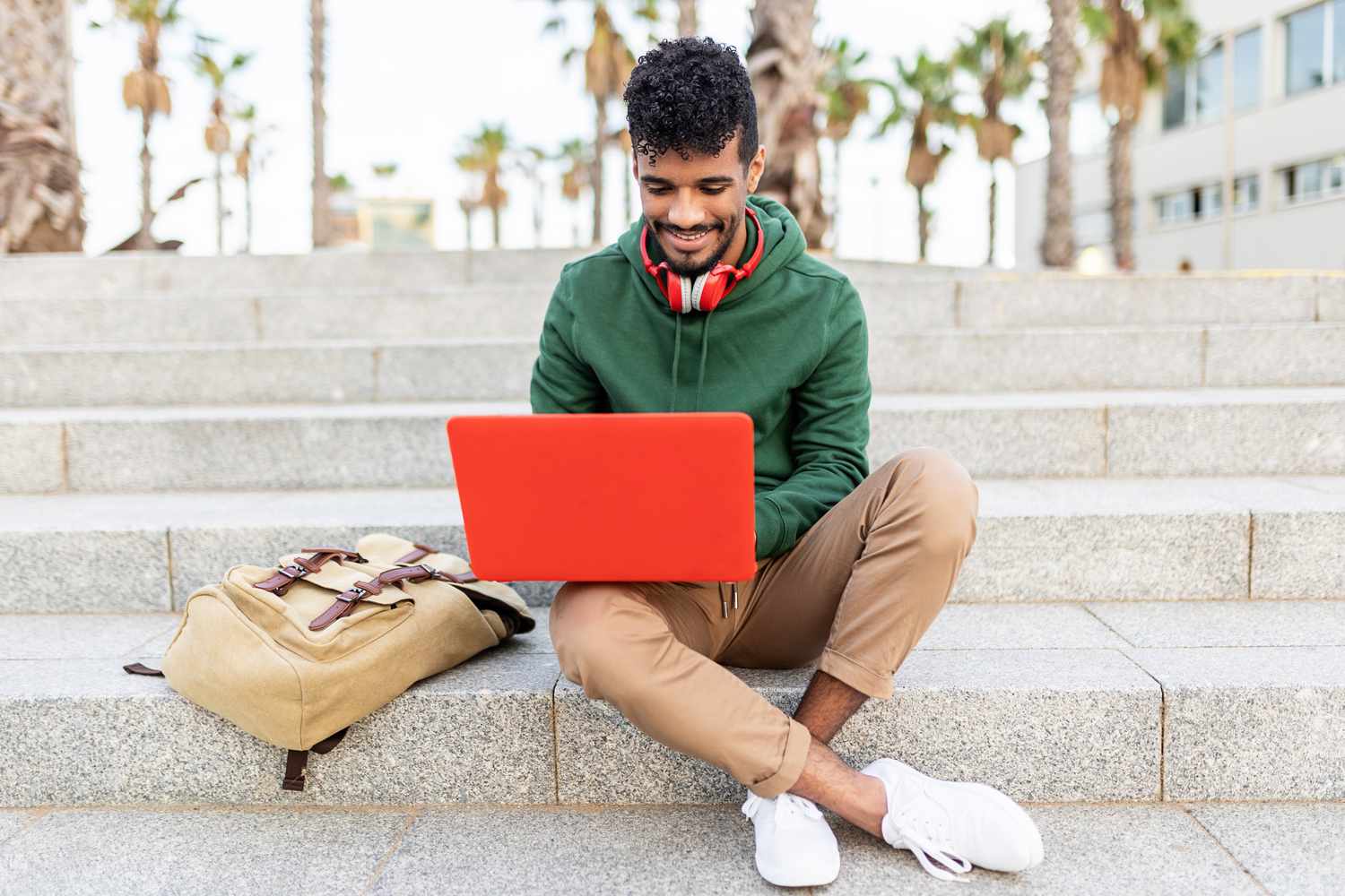 A student sits on the step of a university with a laptop. 