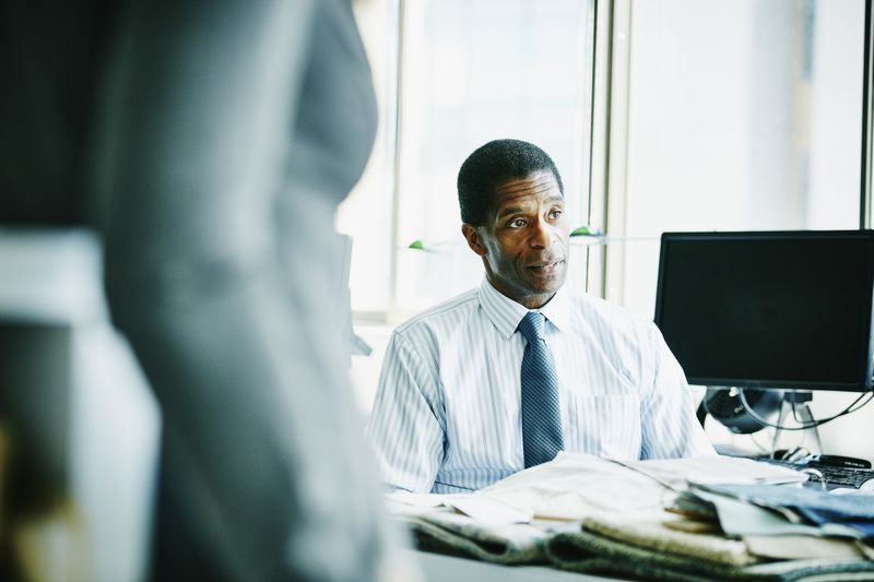 businessman listening with the midsection of another person in the photo and a screen behind him with lots of newspapers and other documents on his desk