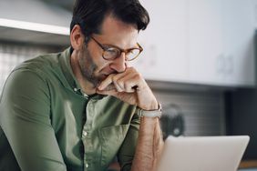 Man in his 40s sitting in his kitchen and seriously pondering financial information on his laptop