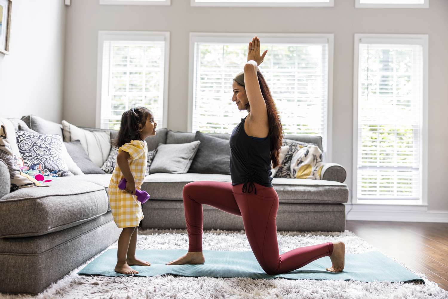 A mother does yoga in the living room while her young daughter stands near holding hand weights