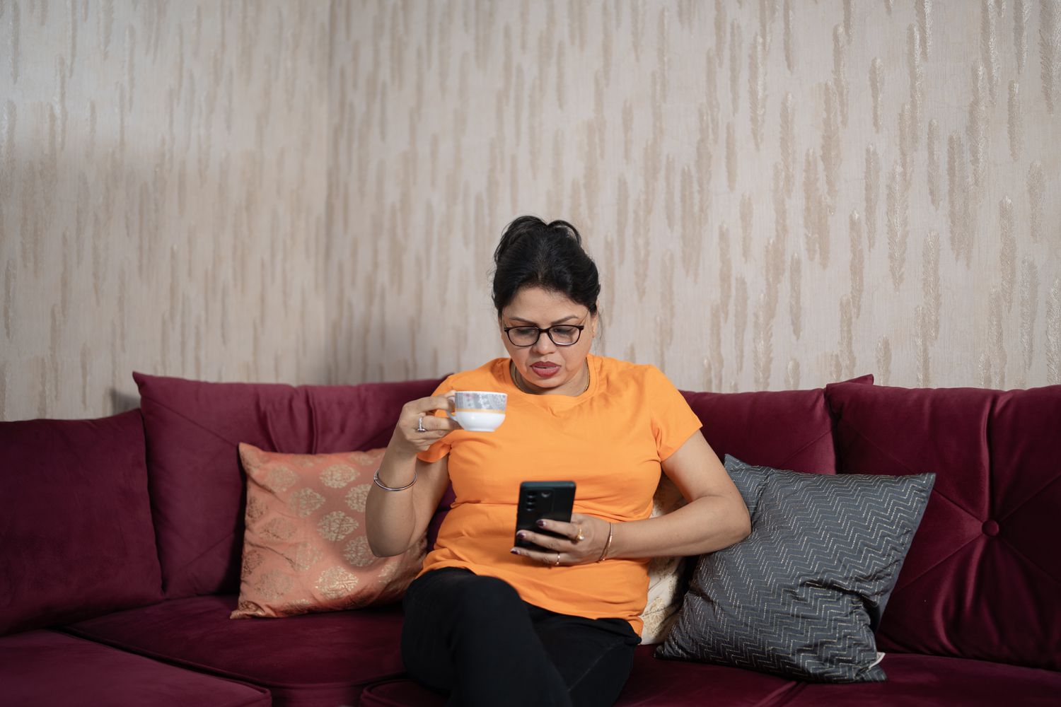 Woman sitting on a couch, holding a cup of tea and studying data on her mobile phone.