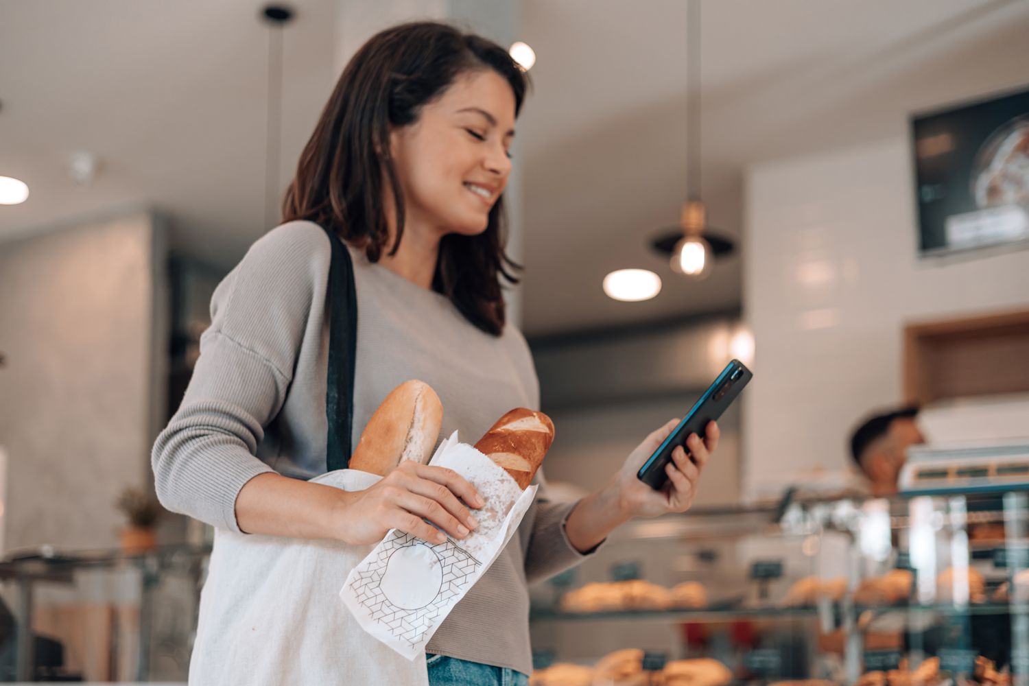 Woman holding a small loaf of bread in a pastery shop. She is looking at her phone. 