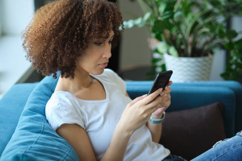Young woman sitting on her sofa and looking seriously at Fed news on her smartphone