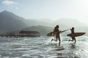 People running on a beach with surfboards with mountains in the background.