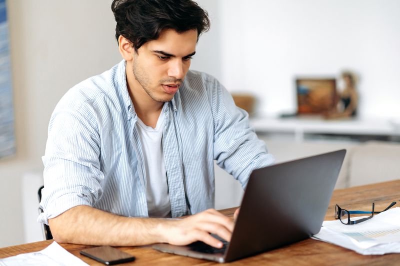 Young man sitting at his home desk and looking seriously at financial information on a laptop