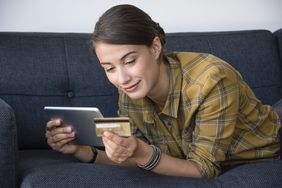 A smiling woman lays on a couch, looking at a bank card and a tablet.