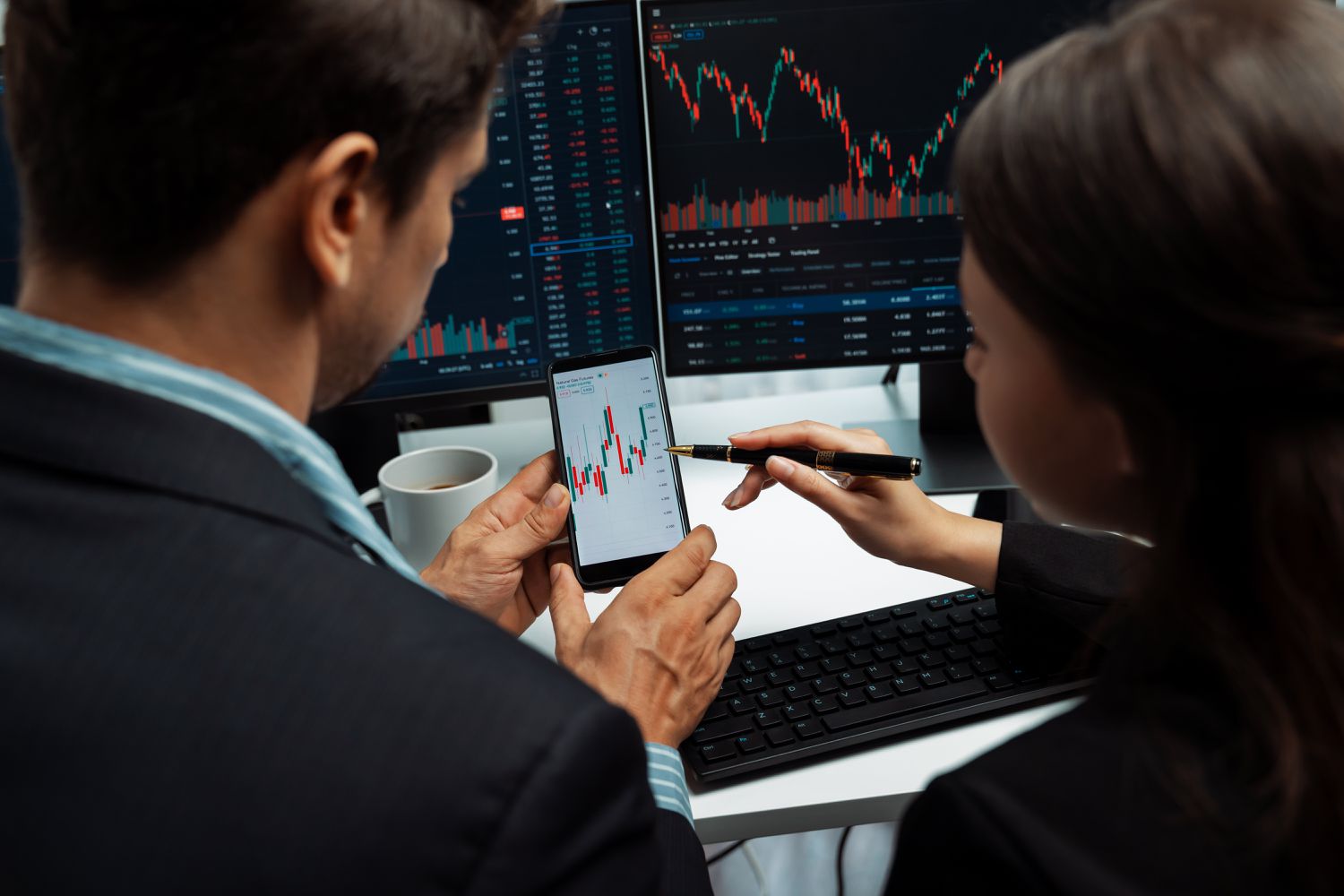 Two traders focusing on a stock chart displayed on phone screen to identify zone of resistance with other information showing on PC screens.