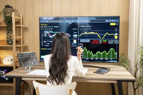 Back of woman showing her sitting at her desk looking at data on multiple monitors.