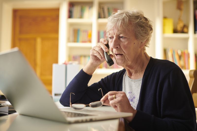An older person on a phone while sitting in front of a laptop