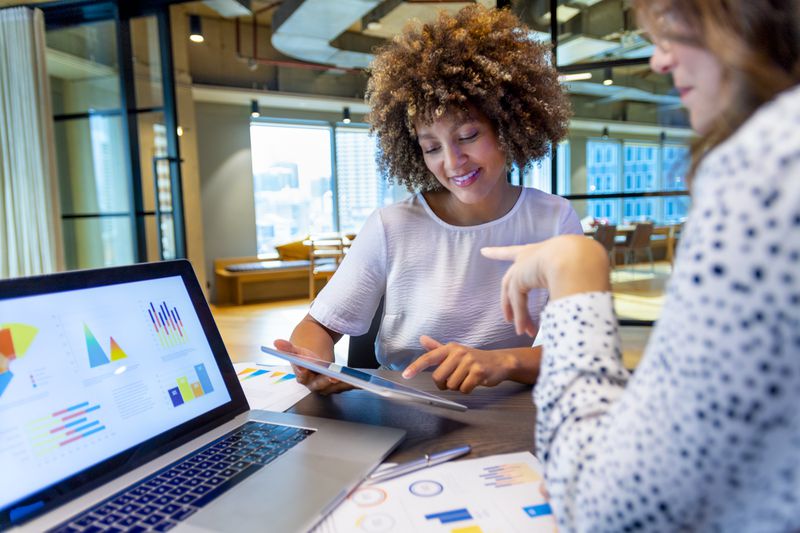 Two colleagues discuss charts on their computers at a work table