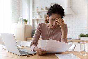 A worried woman looks at paper bills at a table, with a laptop computer.
