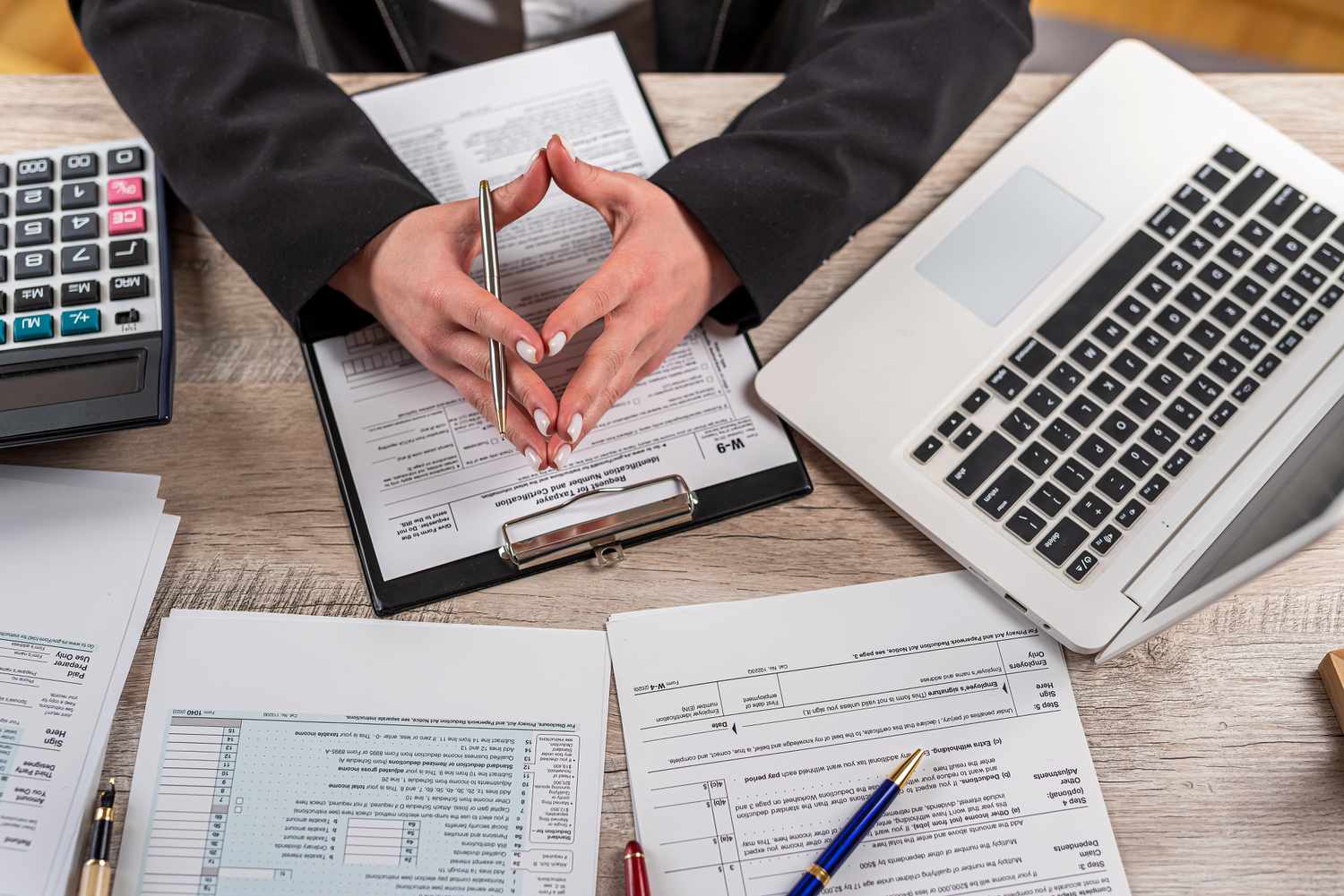 A tax accountant has various tax documents, a calculator, and a laptop spread on their desktop as they calculate taxes for a wealthy client.