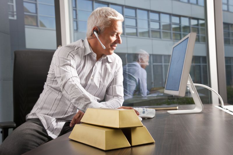 Man sitting at a table working on a computer with gold bars stacked beside him.