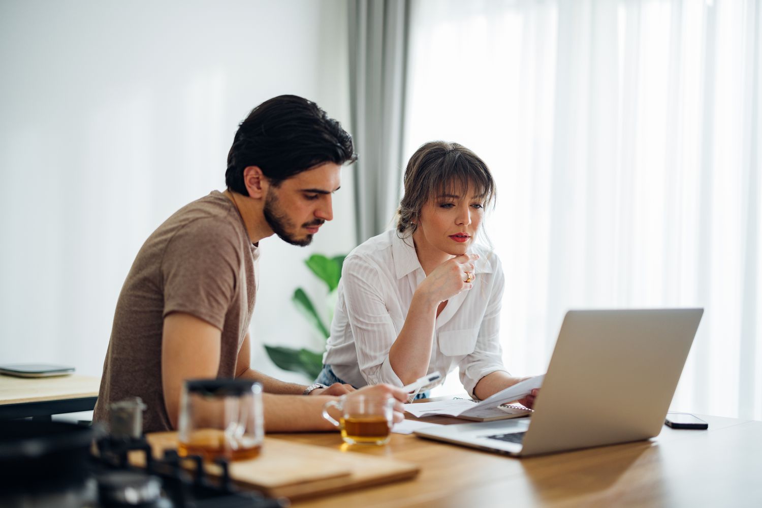 Young couple sitting at kitchen table together and looking serious at a laptop and documents