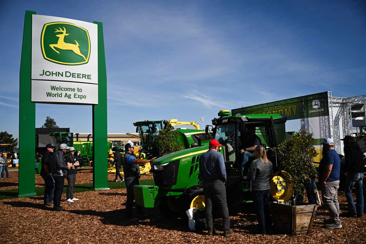 The John Deere logo is seen on a sign above people looking at a green and yellow tractor.