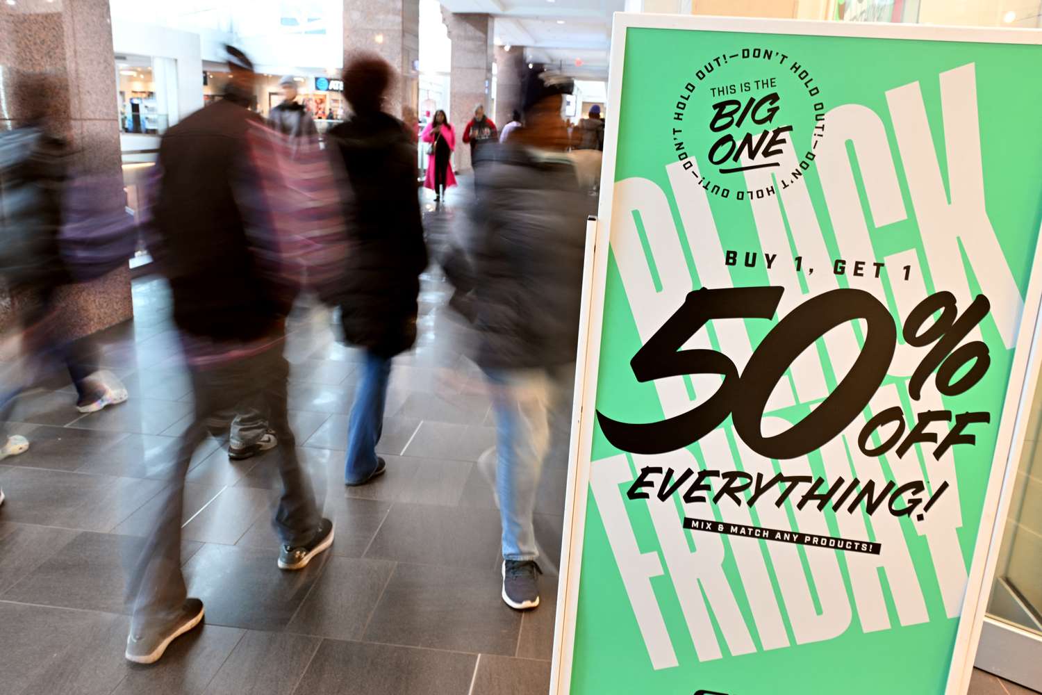 People walk in a mall next to a large green sign that says "Black Friday, Buy 1 Get 1 50% Off Everything!"