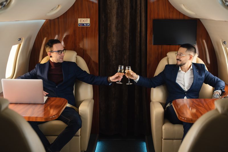 Two well-dressed adults sitting across from each other on a private airplane doing a cheers notion with champagne. 