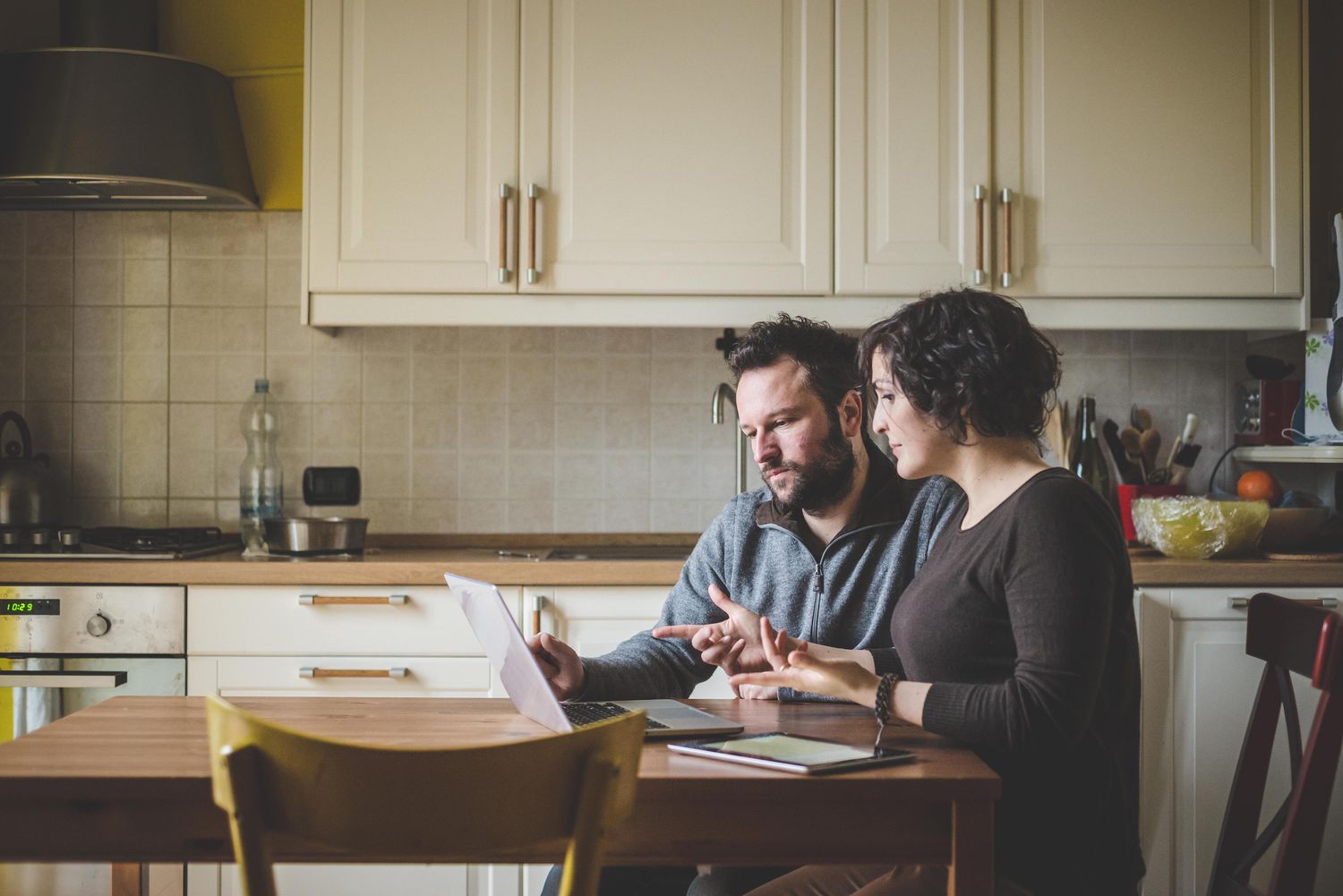 A man and a woman sit at a kitchen table looking at a laptop
