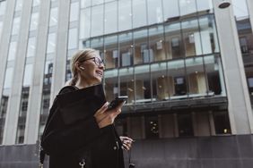 Businesswoman walking in the city surrounded by high-rises, with in-ear headphones on.